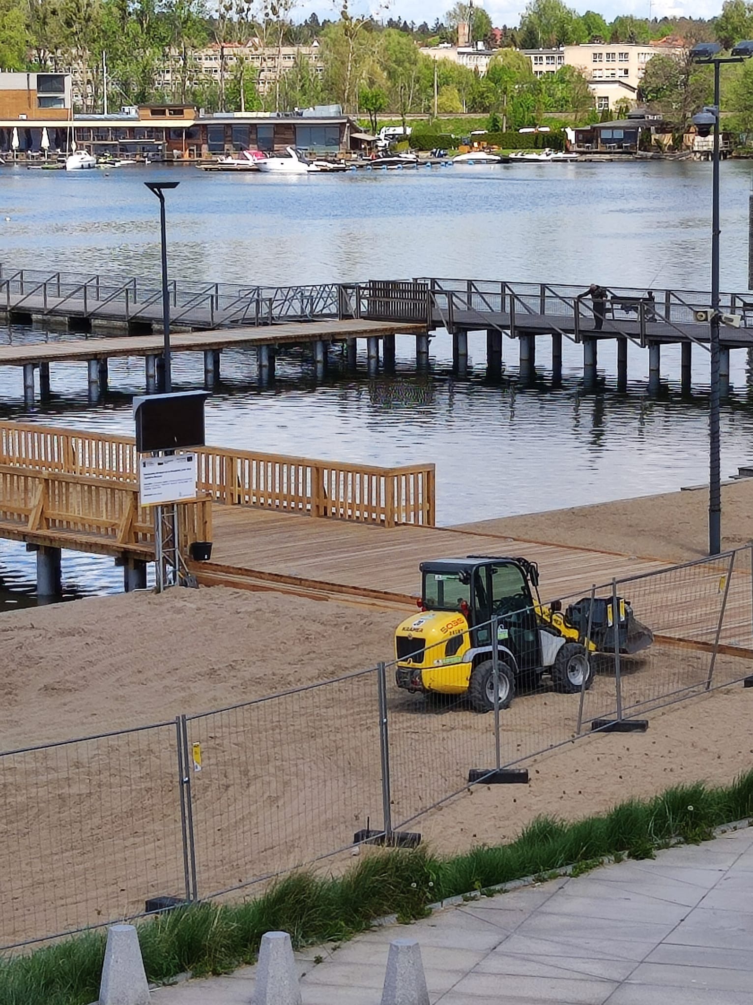 Żółta mini ładowarka Kramer 5055 na piaszczystym terenie budowy pomostu nad jeziorem; w tle widoczne drewniane pomosty, budynki nad brzegiem jeziora i zieleń.