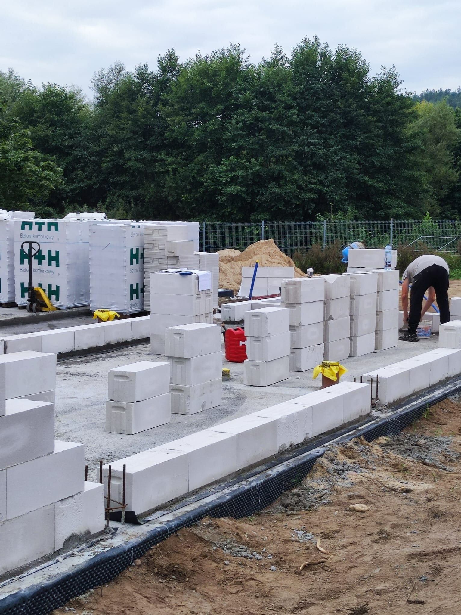 Budowa fundamentów z bloczków betonowych komórkowych na placu budowy, widoczny pracownik przygotowujący zaprawę, palety z bloczkami, izolacja fundamentów i zbrojenie.