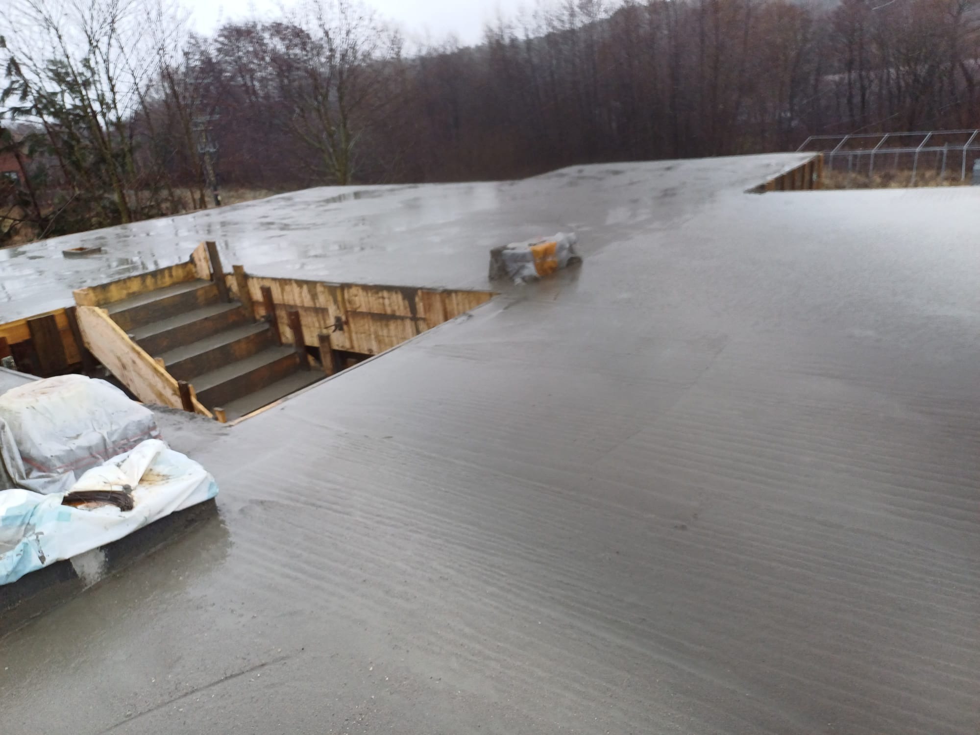 Świeżo wylany beton na tarasie z drewnianym szalunkiem schodów, widoczne worki zabezpieczające przed wysychaniem, tło stanowią drzewa bez liści.