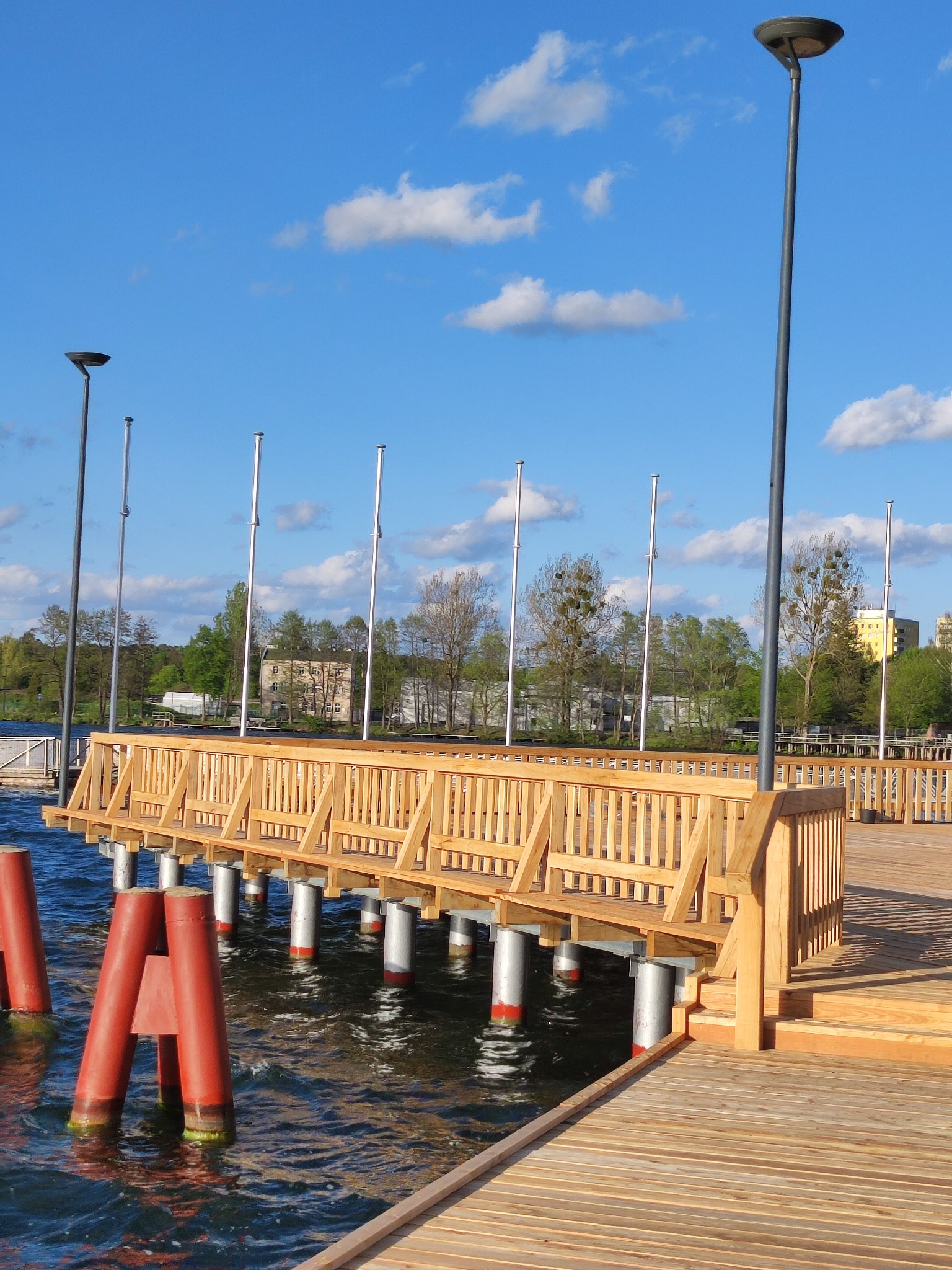 Nowoczesny drewniany pomost nad wodą z metalowymi podporami, oświetlony wysokimi lampami, z widokiem na budynki i drzewa w tle pod błękitnym niebem z chmurami.