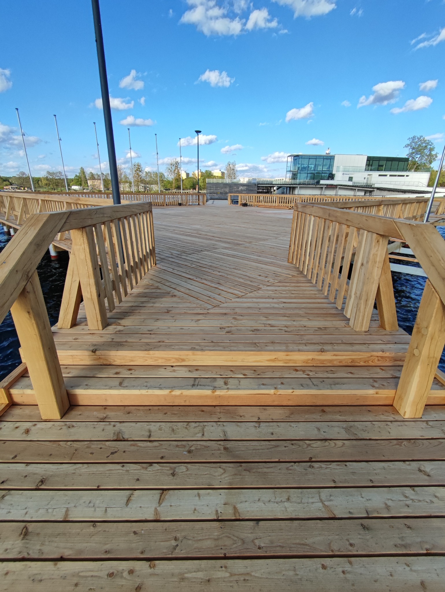 Nowy drewniany taras z jasnego drewna, z widocznymi schodami i balustradami, prowadzący w kierunku wody i nowoczesnego budynku w tle, pod błękitnym niebem z białymi chmurami.