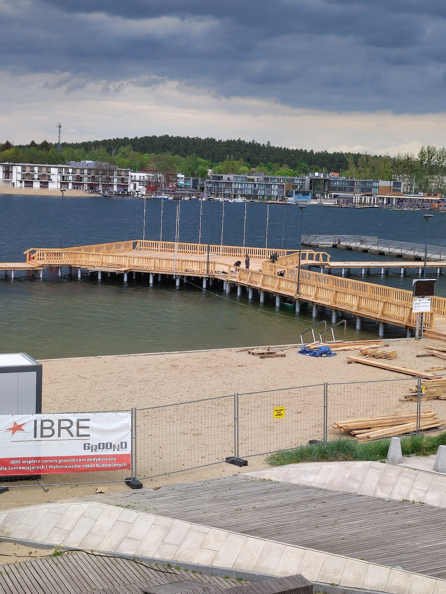 Budowa drewnianego pomostu nad jeziorem w Pszczółkach, widok z góry na konstrukcję w trakcie montażu, z widocznymi elementami rusztowania i przygotowanymi deskami.
