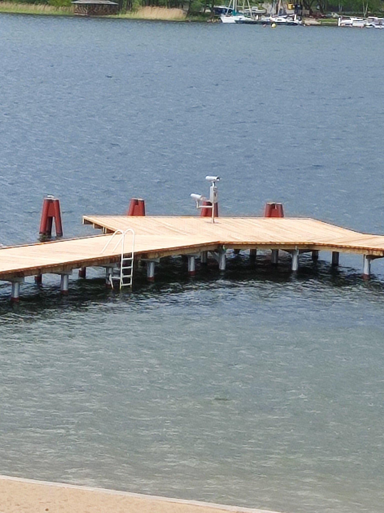 Nowy drewniany pomost z drabinką do wody, wsparty na betonowych filarach, z czerwonymi słupkami i kamerą monitoringu, widok z plaży na jezioro.