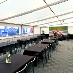 Motorsport Location Catering Ltd - Ustawione stoły z czarnymi obrusami i szarymi krzesłami w białym namiocie, dekoracje kwiatowe na stołach, w tle baner z motocyklem wyścigowym i rośliny doniczkowe.