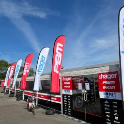 Motorsport Location Catering Ltd - Stoisko promocyjne z flagami i logotypami sponsorów, w Krakowie, przed wejściem zaparkowany skuter.