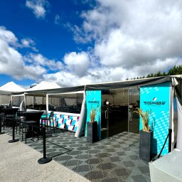 Motorsport Location Catering Ltd - Zadaszone namioty z logo EGMBE Motorsport, czarne krzesła i stoliki na zewnątrz, podłoga z szarej kostki, niebieskie niebo z chmurami