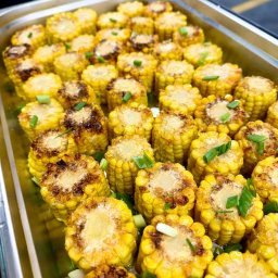 Motorsport Location Catering Ltd - Blacha z równo ułożonymi, grillowanymi kolbami kukurydzy, każda udekorowana posiekanym szczypiorkiem, widoczna lekka glazura i miejscami przypalenia.