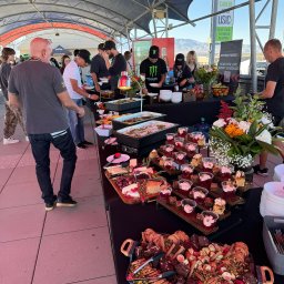 Motorsport Location Catering Ltd - Bufet z różnorodnymi potrawami: ciepłe dania w podgrzewaczach, desery w plastikowych kubeczkach, sery i owoce na drewnianych deskach, ciasta na paterach, kwiatowe dekoracje, obsługa w czarnych...