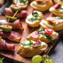 Motorsport Location Catering Ltd - Drewniana deska z różnorodnymi, apetycznymi przekąskami: figi owinięte szynką parmeńską na wykałaczkach, grzanki z kremowym serem i figami, grzanki z pomidorkami i rukolą, całość uzupełniona...