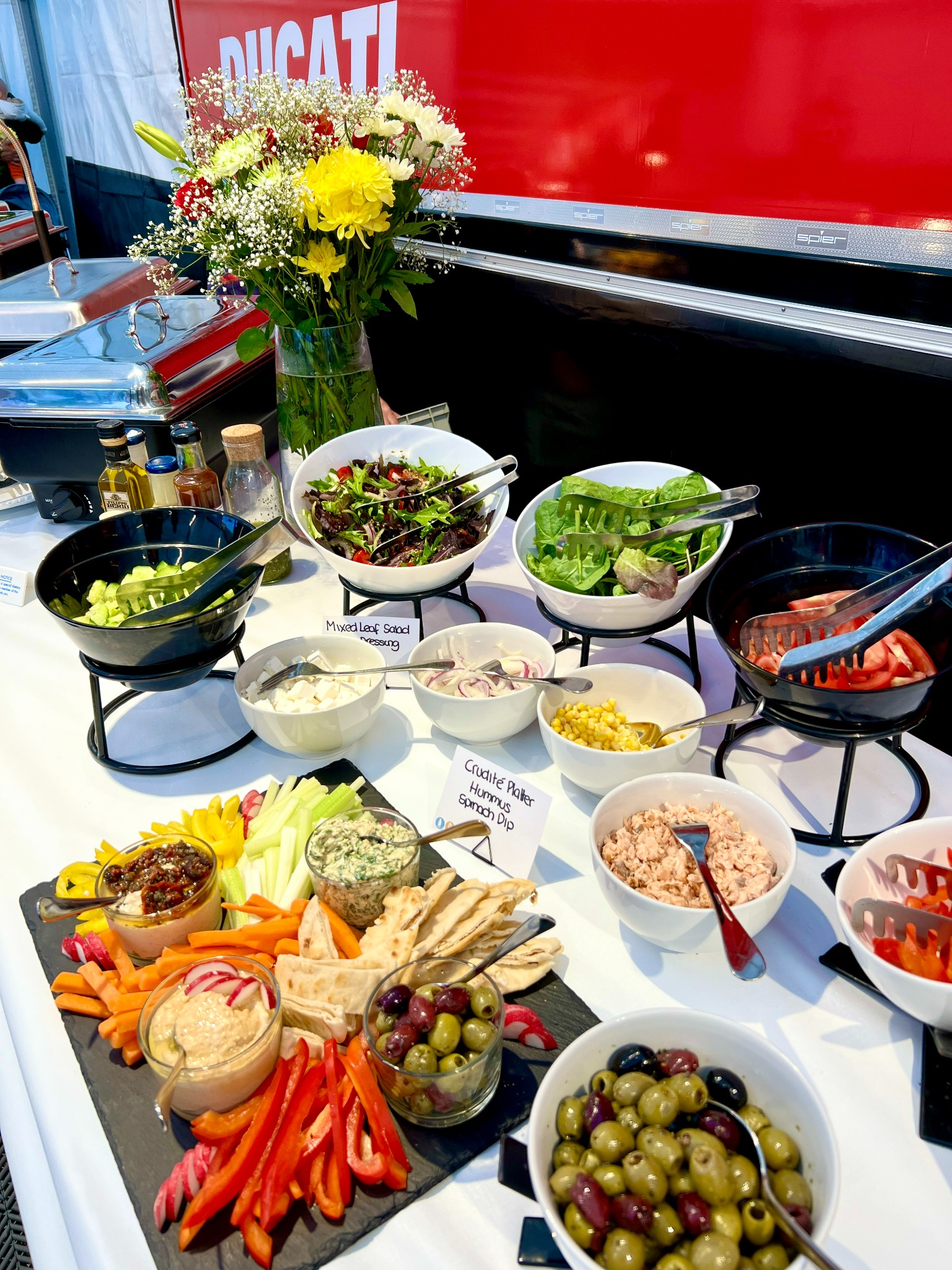 Stół z różnorodnymi przekąskami: sałatki, warzywa, hummus, oliwki, patery z dipami, kwiaty w wazonie, ekspozycja dań na białym obrusie z logo Ducati w tle.