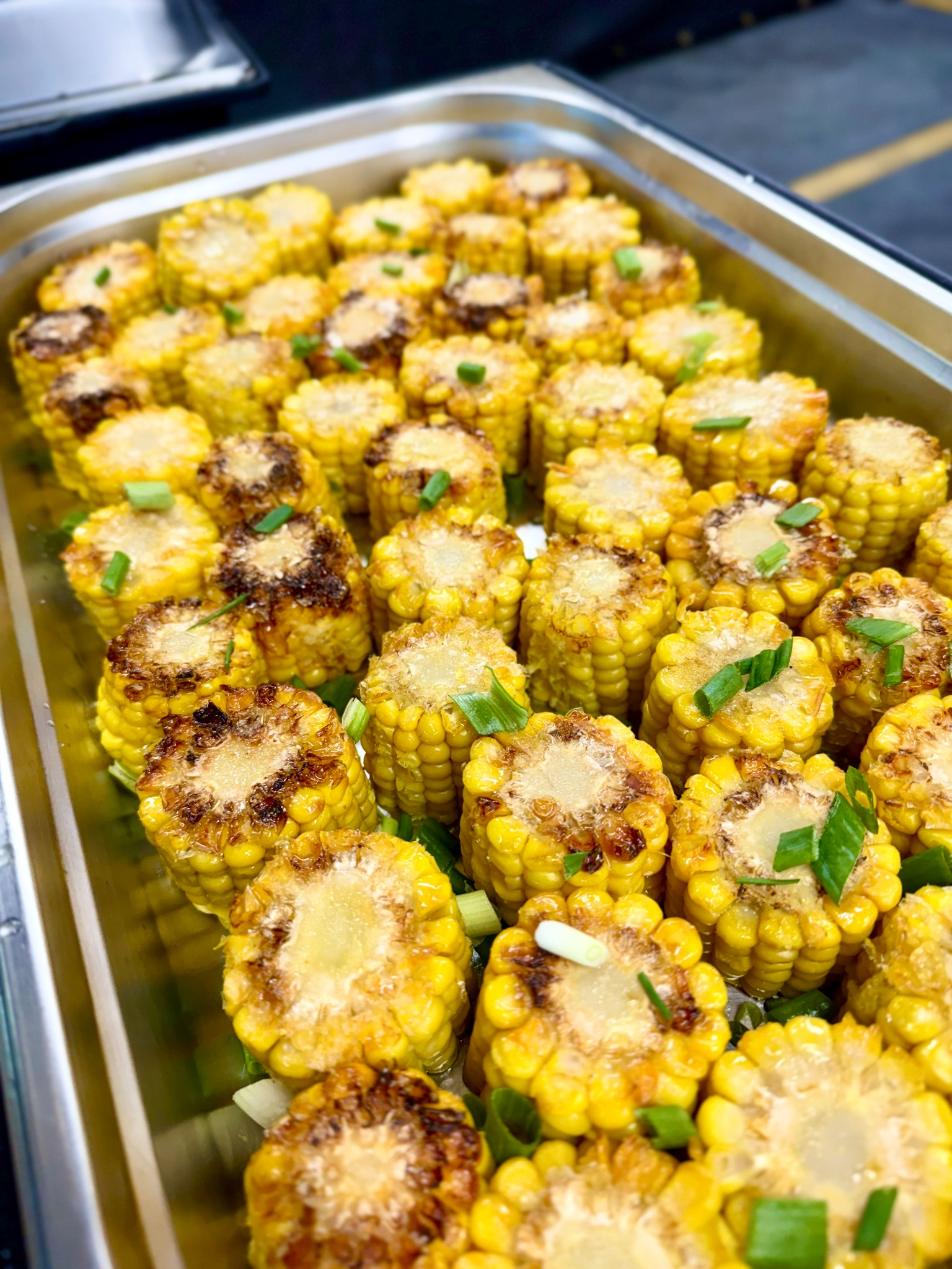 Blacha z równo ułożonymi, grillowanymi kolbami kukurydzy, każda udekorowana posiekanym szczypiorkiem, widoczna lekka glazura i miejscami przypalenia.