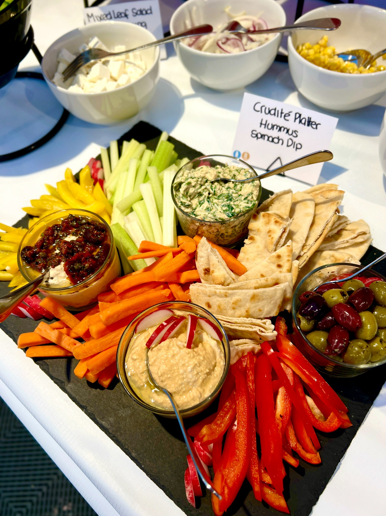 Elegancko podany talerz crudités na czarnym łupku z różnymi dipami, warzywami w słupkach, oliwkami i pieczywem pita; dodatkowo miseczki z sałatkami w tle.
