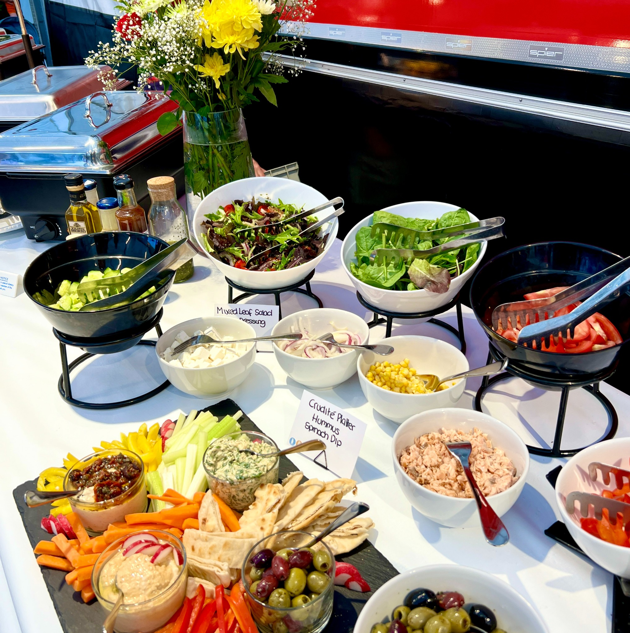 Bufet sałatkowy z różnymi składnikami, w tym sałatami mieszanymi, warzywami, dipami i oliwkami, umieszczony na białym stole z dekoracjami kwiatowymi w tle.