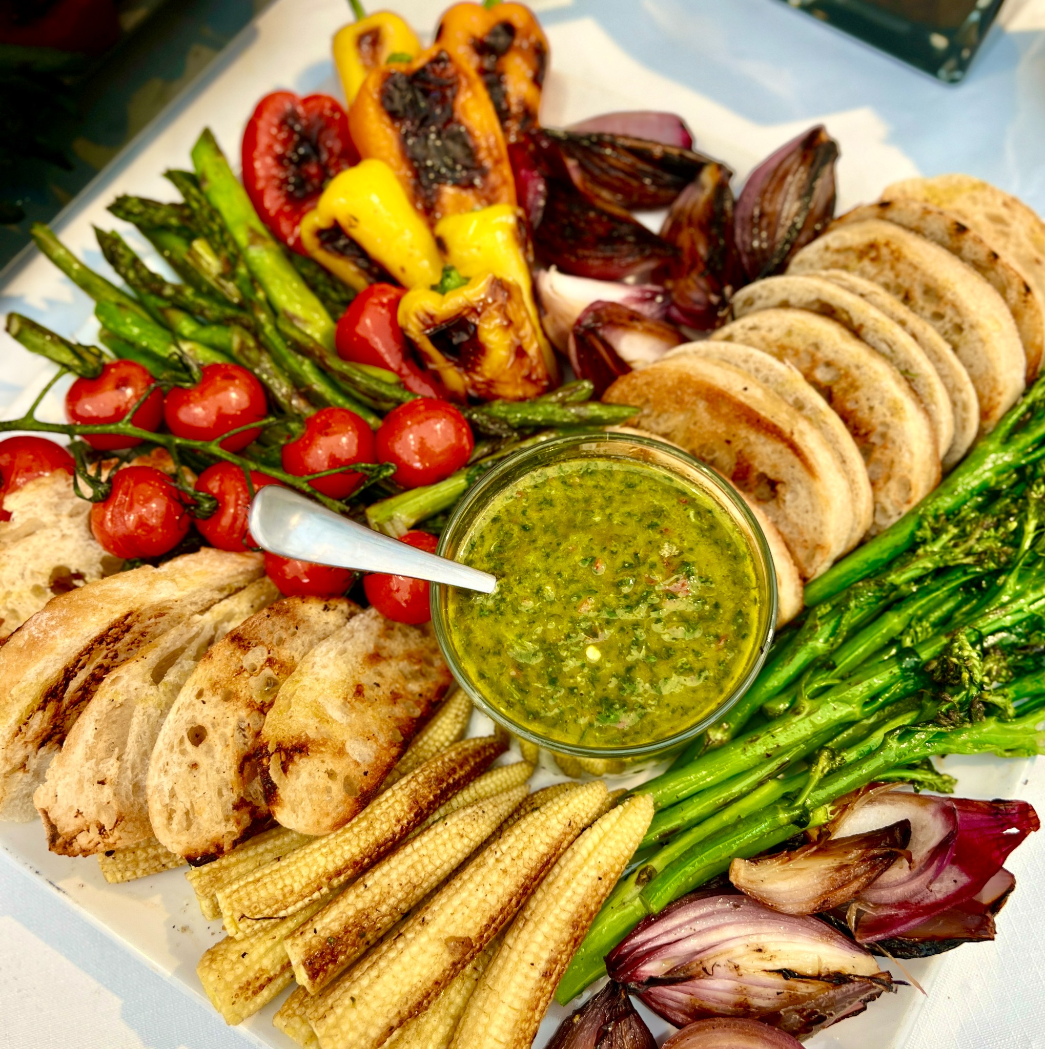 Elegancko ułożona na białym talerzu kompozycja grillowanych warzyw: papryki, cebuli, szparagów, pomidorków koktajlowych na gałązkach, mini kukurydzy, brokułów, pieczywa i miseczki z zielonym sosem.