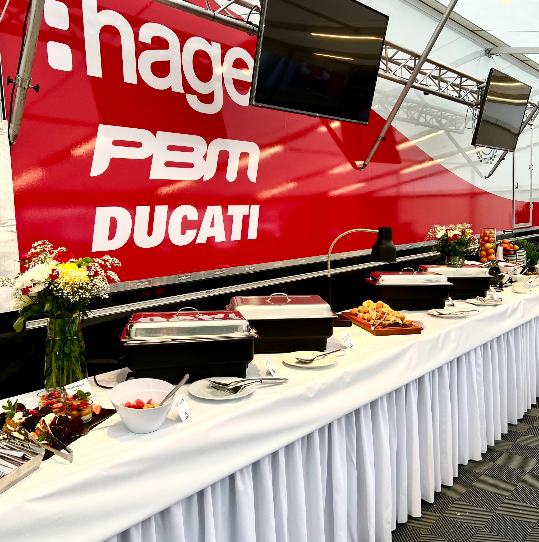 Bufet z przekąskami i deserami na białym obrusie, w tle czerwony baner z logo i dwa ekrany LCD.