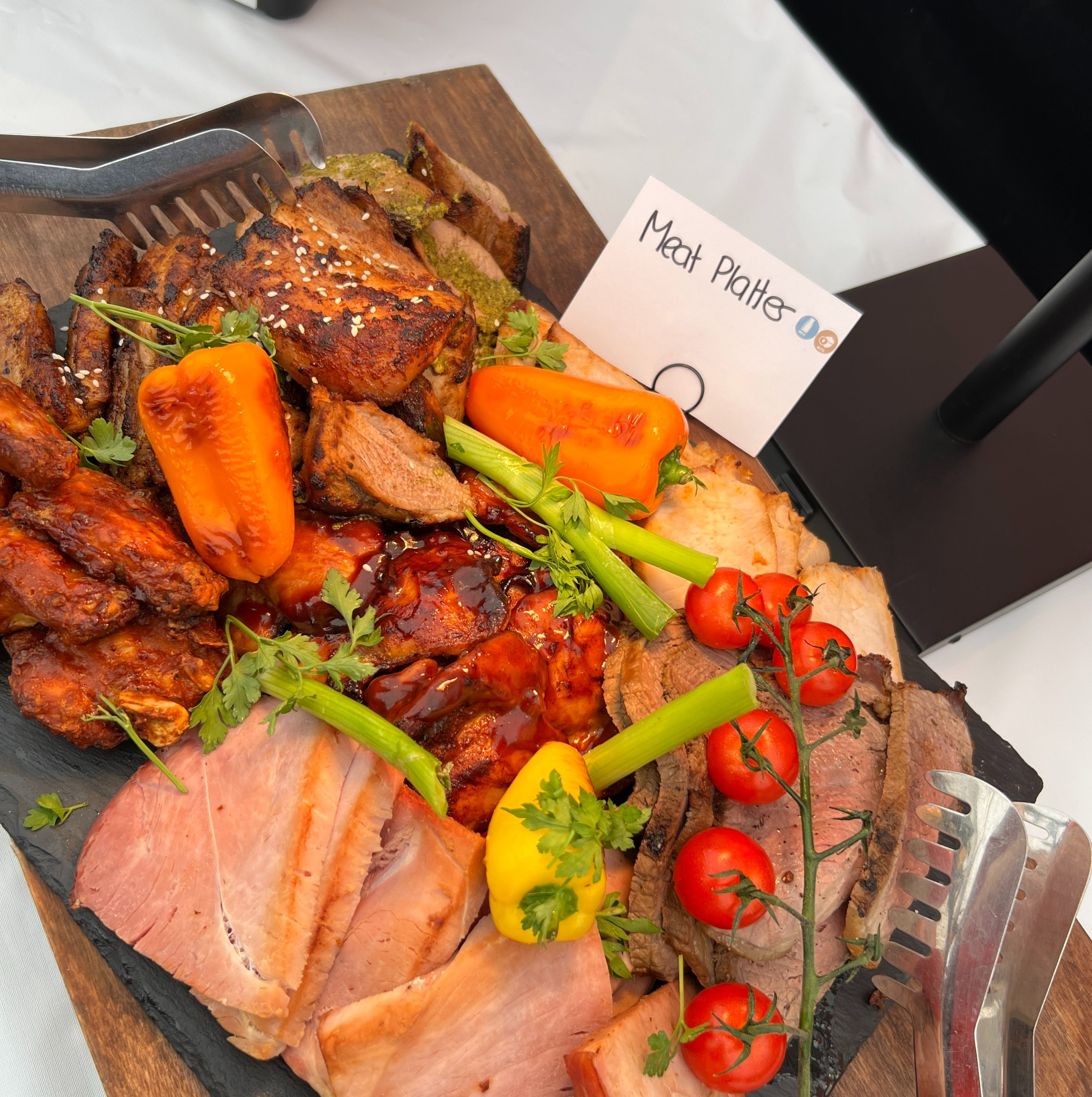 Bogata deska mięs: pieczone żeberka z sezamem, skrzydełka w sosie BBQ, plasterki szynki i pieczeni wołowej, dekorowane papryką, selerem naciowym, pomidorkami cherry i natką pietruszki, z kartką...