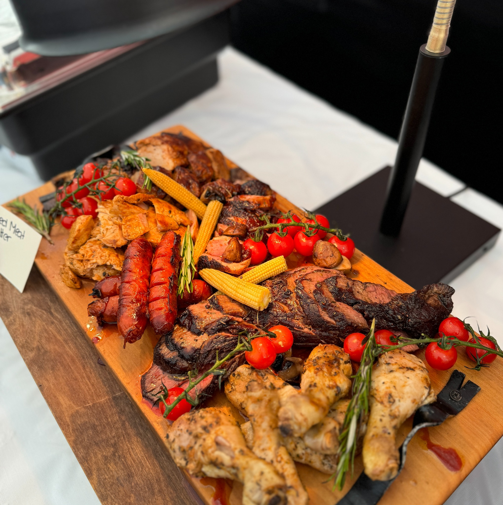 Deska z grillowanymi mięsami: kurczak, wołowina, kiełbaski, kukurydza i pomidorki cherry, udekorowana gałązkami rozmarynu, z widoczną kartką z napisem 'Mixed Meat Platter'.