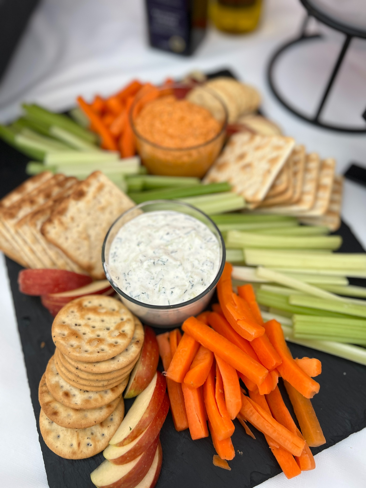 Elegancko ułożony talerz z warzywami, krakersami i dipami na czarnym tle, z marchewką, selerem naciowym, jabłkami i dwoma rodzajami dipów w szklanych miseczkach.