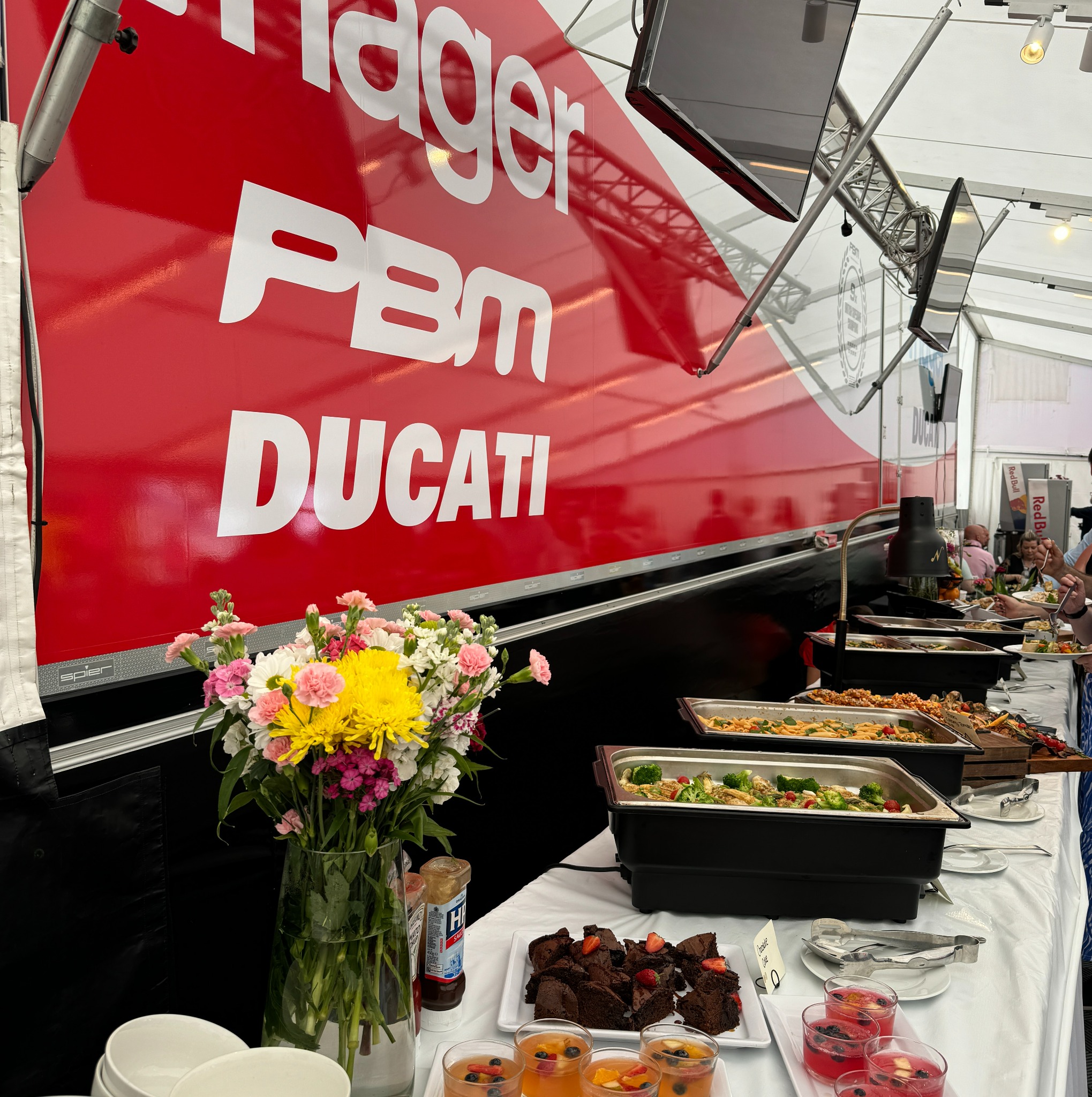 Bufet z różnorodnymi daniami, w tym makaronami, warzywami, brownie i galaretkami owocowymi, udekorowany bukietem kwiatów na tle czerwonego baneru z logo PBM Ducati.