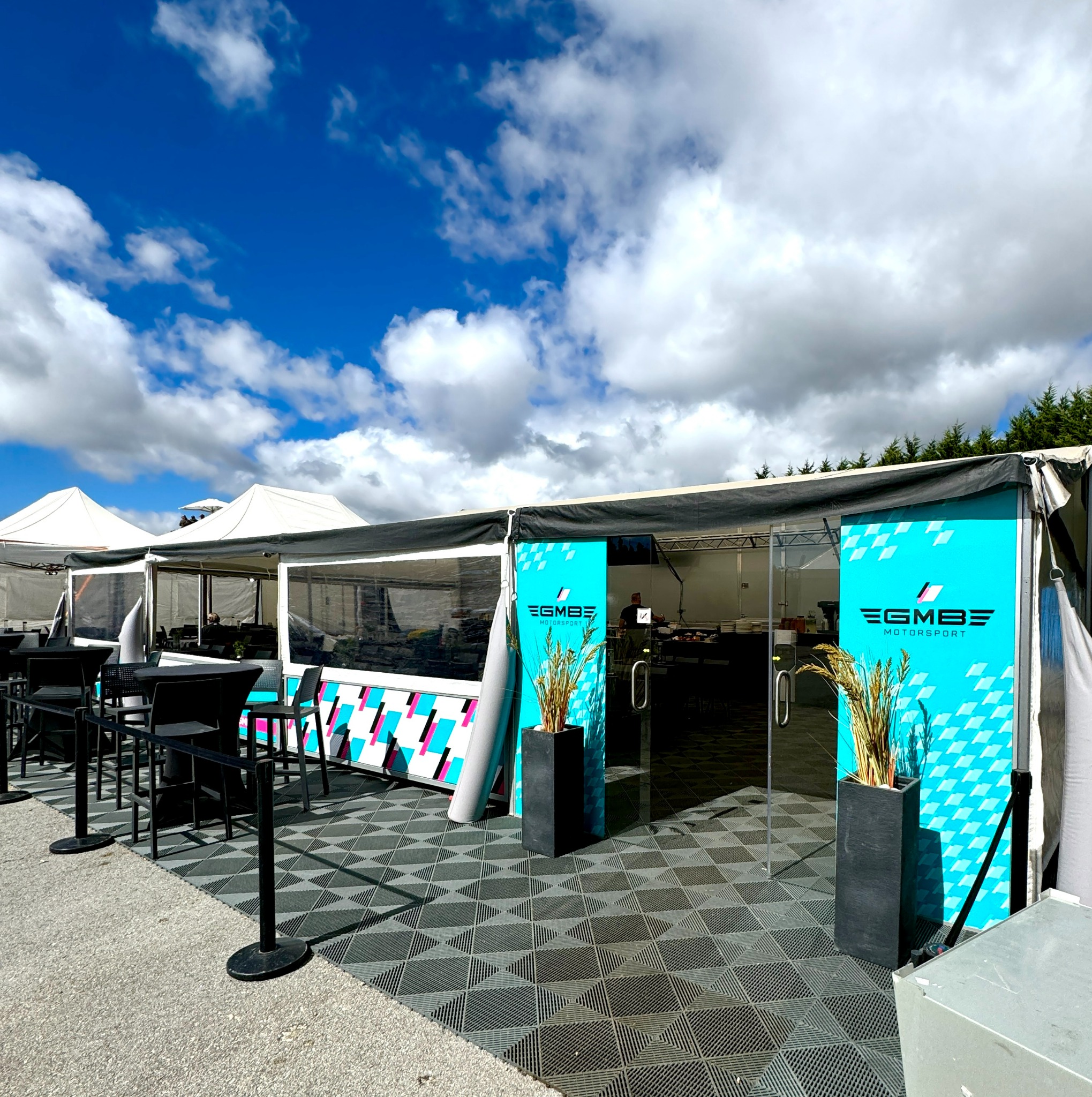 Namiot imprezowy z logo EGMBE Motorsport i dekoracyjnymi trawami w donicach, z widocznymi stolikami i krzesłami oraz pracownikiem wewnątrz.
