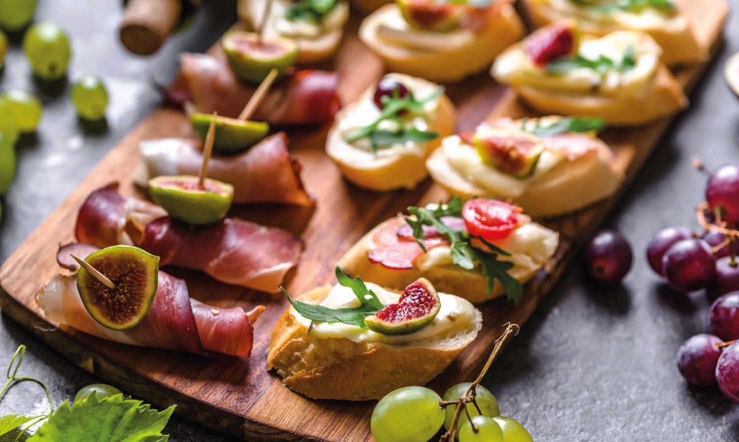 Drewniana deska z różnorodnymi, apetycznymi przekąskami: figi owinięte szynką parmeńską na wykałaczkach, grzanki z kremowym serem i figami, grzanki z pomidorkami i rukolą, całość uzupełniona...