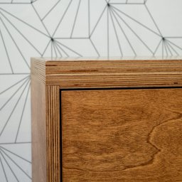 PRACOWNIA STOLARSKA MARCIN ŻEBROWSKI - Szafka ze sklejki wykończona bejcą i olejowoskiem, widoczny detal krawędzi. Realizacja stolarza z Łodzi. Geometryczna tapeta w tle.