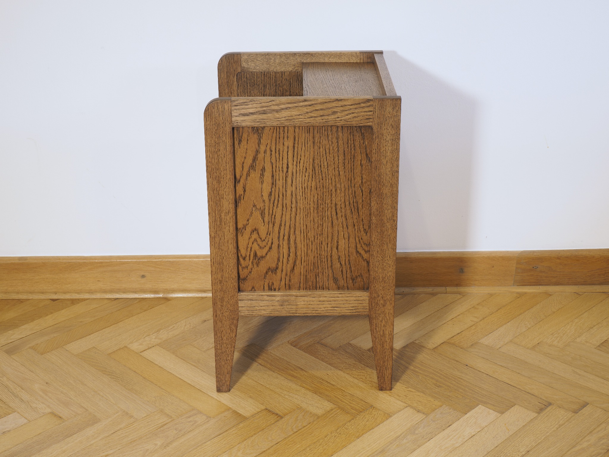 Dębowa szafka nocna z widocznym usłojeniem drewna, ustawiona na parkiecie w jodełkę, na tle białej ściany. Minimalistyczny design.