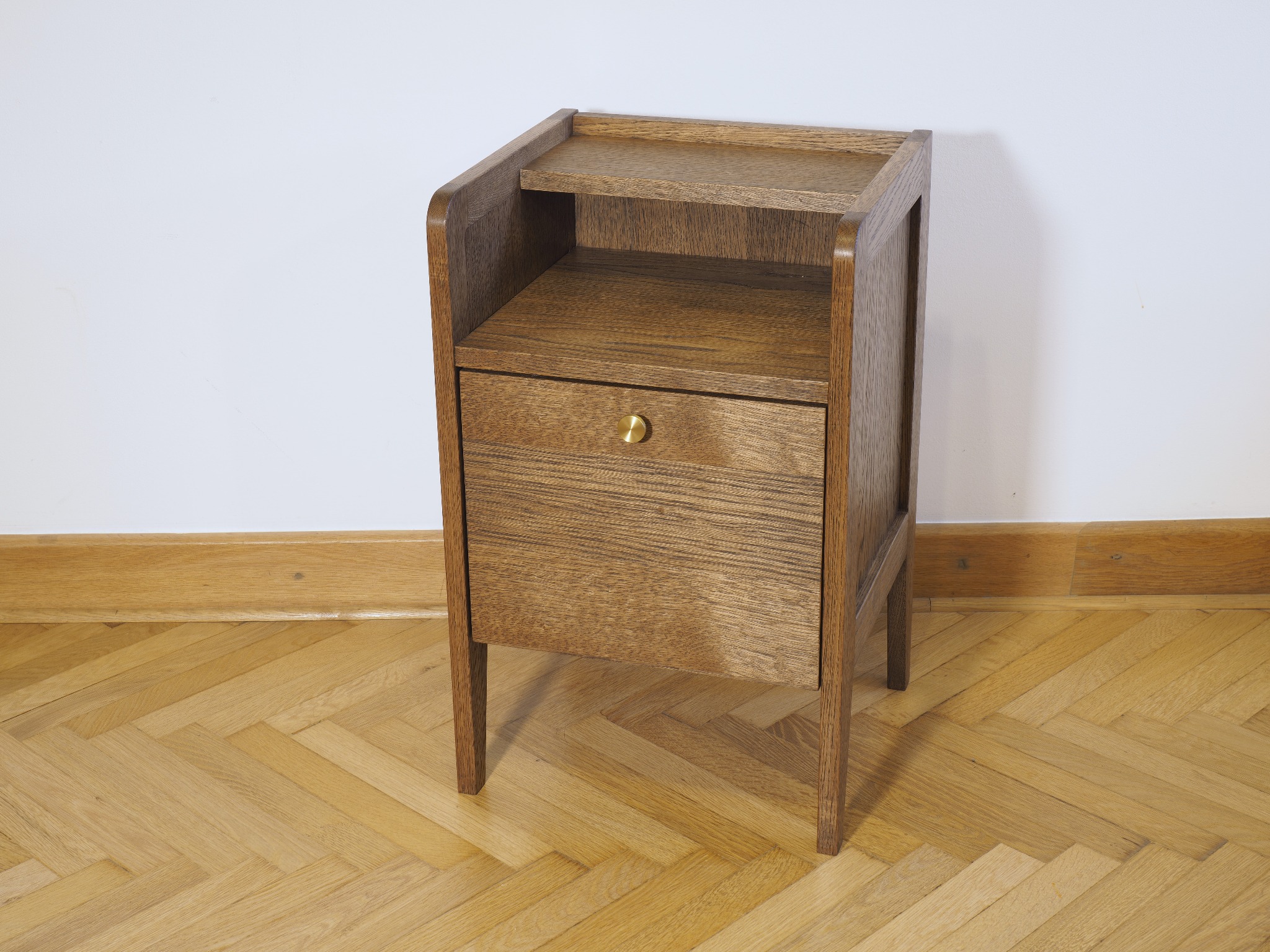 Dębowa szafka nocna z szufladą i półką, widoczna na parkiecie w jodełkę, z bliska na tle białej ściany. Mosiężny uchwyt szuflady.