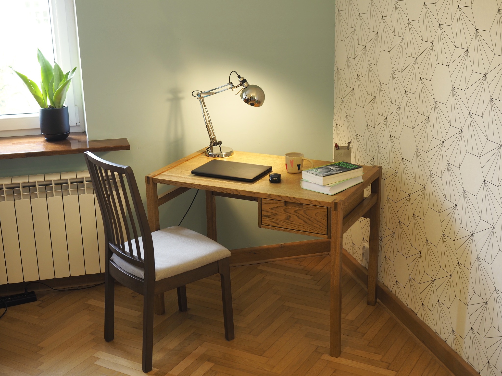 Dębowe biurko z szufladą, wykończone bejcą olejną i woskiem. Na blacie lampka, laptop, książki. Krzesło z szarą tapicerką. W tle ściana w geometryczne wzory.