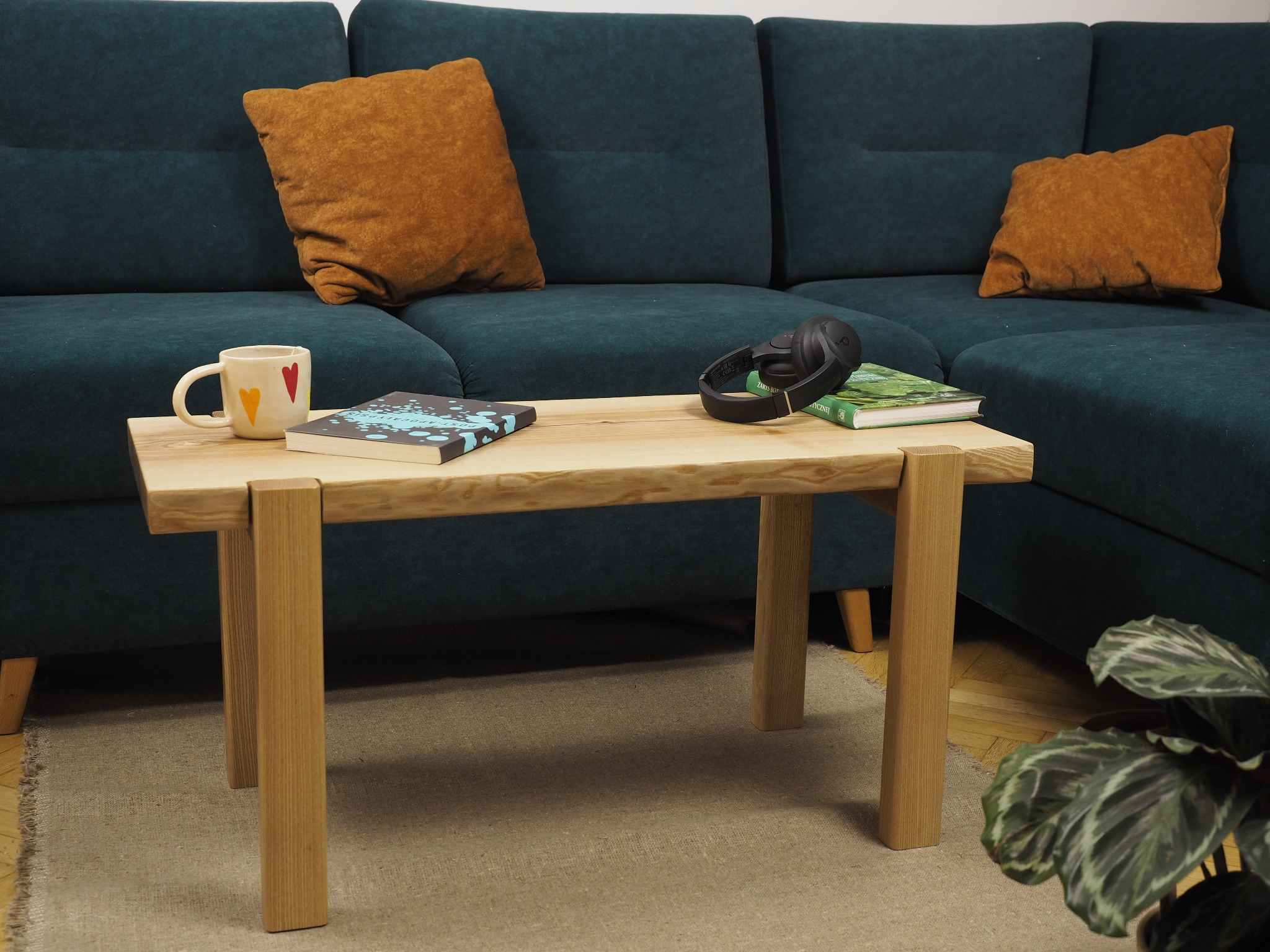 Stolik kawowy z jasnego drewna jesionowego z naturalną krawędzią blatu, stojący na beżowym dywanie, z zieloną sofą w tle. Na blacie książki, słuchawki i kubek.