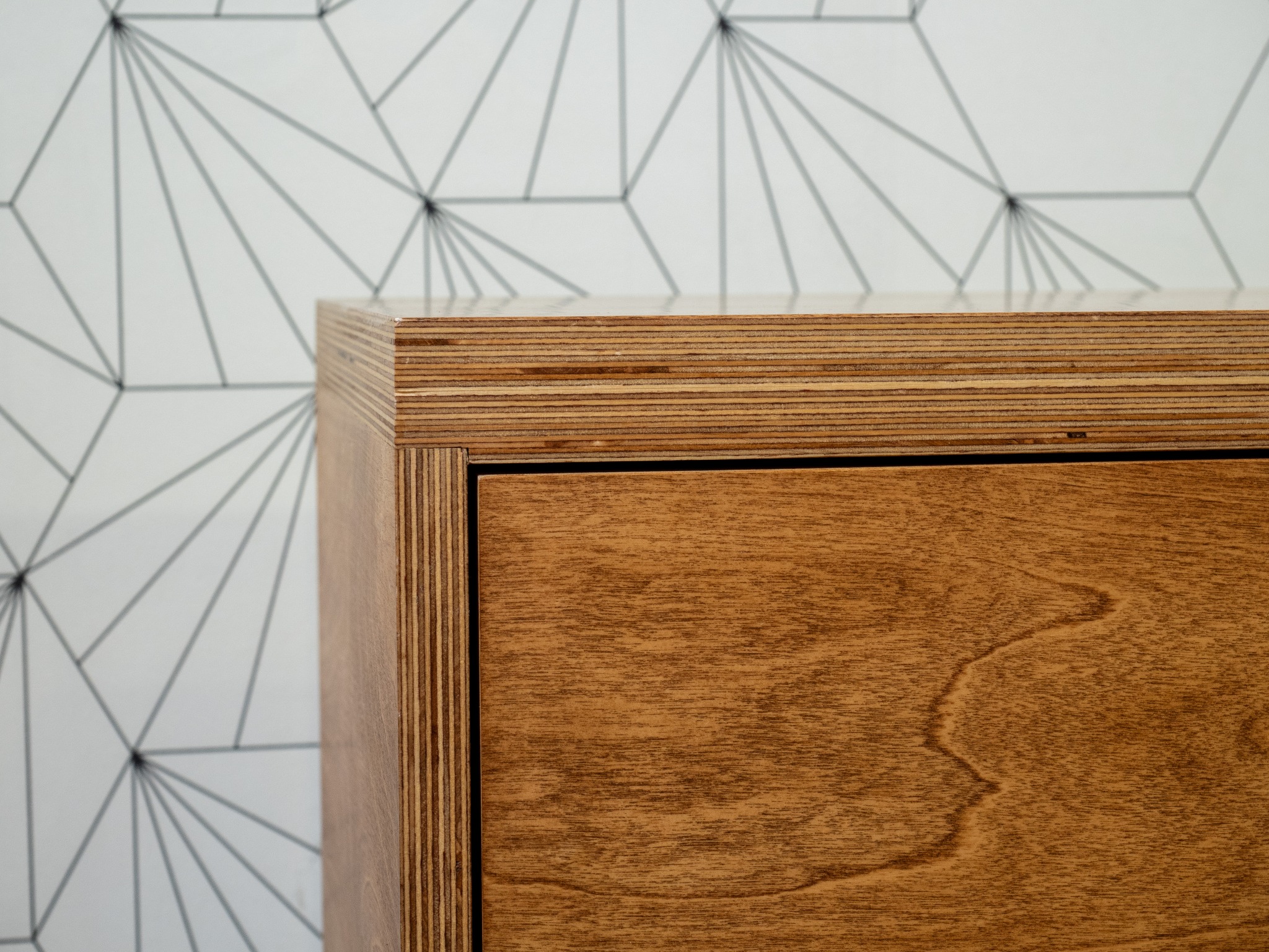 Szafka ze sklejki wykończona bejcą i olejowoskiem, widoczny detal krawędzi. Realizacja stolarza z Łodzi. Geometryczna tapeta w tle.