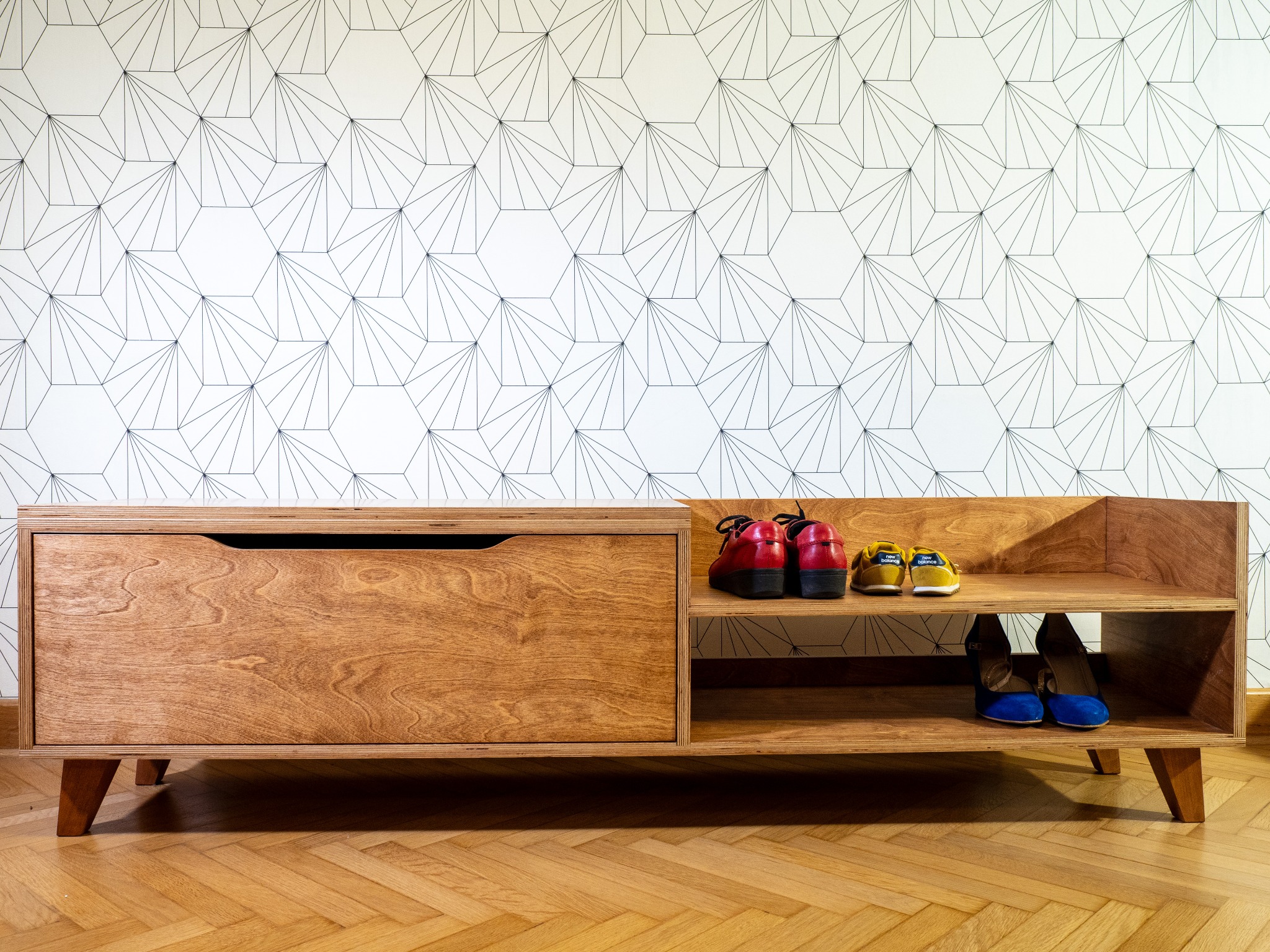 Szafka na buty ze sklejki z siedziskiem, wykończona bejcą olejną i olejowoskiem. Na półkach widoczne buty. Tło stanowi ściana z geometrycznym wzorem.