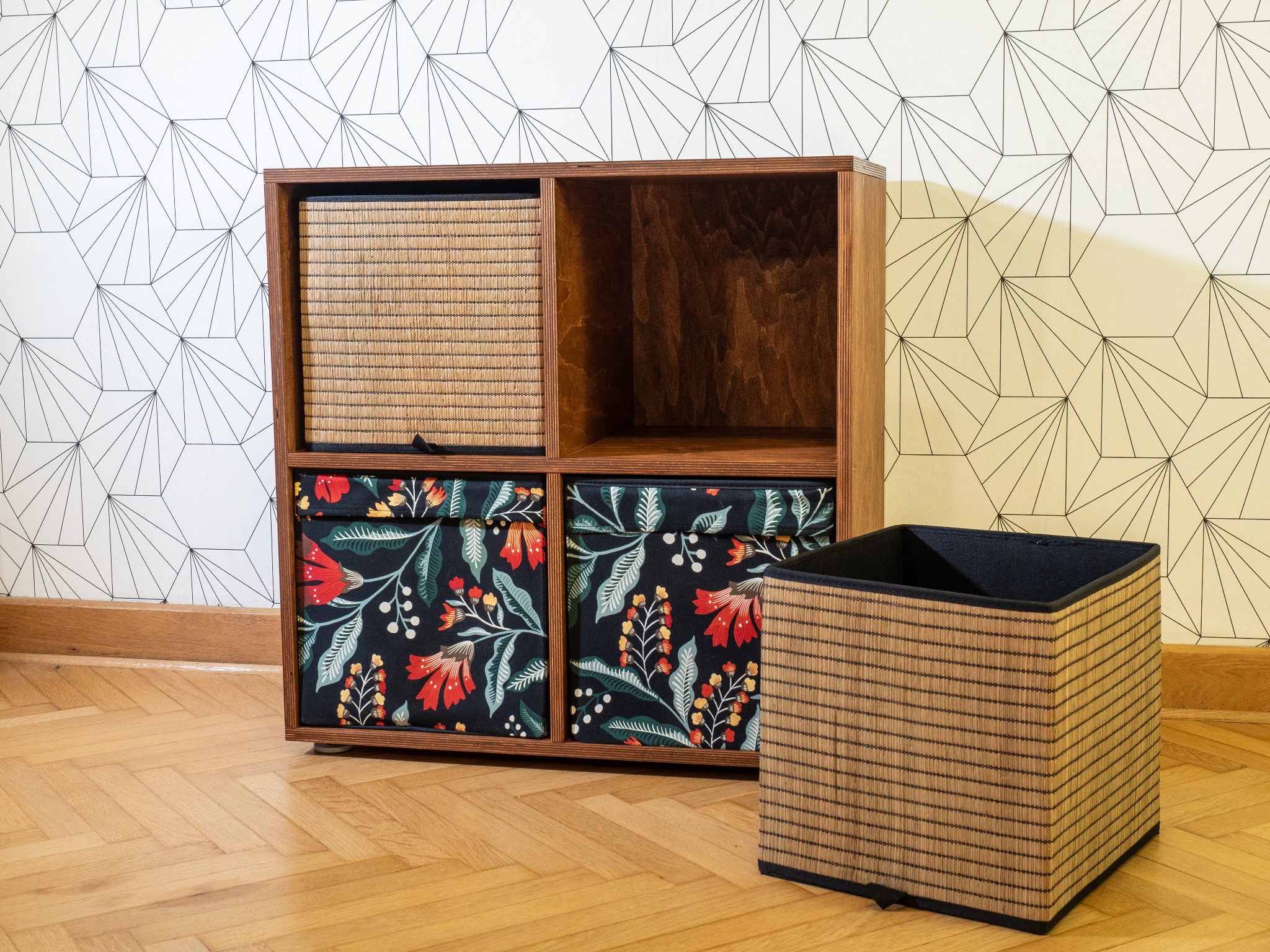 Drewniany regał na pudełka ze sklejki, z dwoma pudełkami w kwiaty i dwoma plecionymi. Na parkiecie, pod białą ścianą w geometryczne wzory.