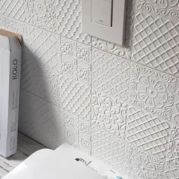 Biała miska WC podwieszana, zamontowana na tle dekoracyjnych, strukturalnych płytek ściennych w łazience, widoczny przycisk spłukujący w kwadratowej ramce i częściowo kartonowe opakowanie.