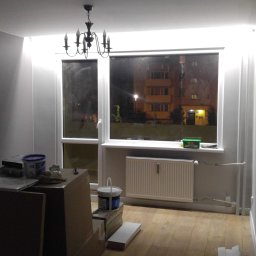 FIRMA HYLEK Jarosław Chylicki - Pokój po malowaniu ścian na szaro, widoczny parkiet, okno z widokiem na oświetlony blok nocą, grzejnik pod oknem, puszki z farbą i narzędzia malarskie