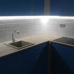 Nowoczesna kuchnia z niebieskimi szafkami, drewnianym blatem, białymi płytkami z delikatnym wzorem i podświetleniem LED nad blatem, z widocznym zlewem w odcieniach szarości i czarną płytą indukcyjną.