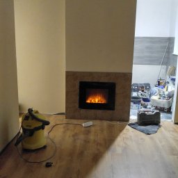 FIRMA HYLEK Jarosław Chylicki - Wnętrze w trakcie prac wykończeniowych: imitacja kominka z brązowymi płytkami, odkurzacz przemysłowy, widoczne narzędzia w tle, świeżo położona podłoga laminowana.