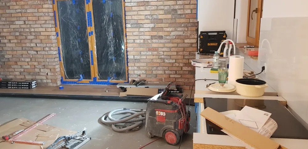 Wnętrze kuchni w trakcie prac wykończeniowych: widoczne zabezpieczone folią okna z ceglaną ścianą, położona podłoga, odkurzacz przemysłowy i narzędzia na podłodze, elementy zabudowy kuchennej...