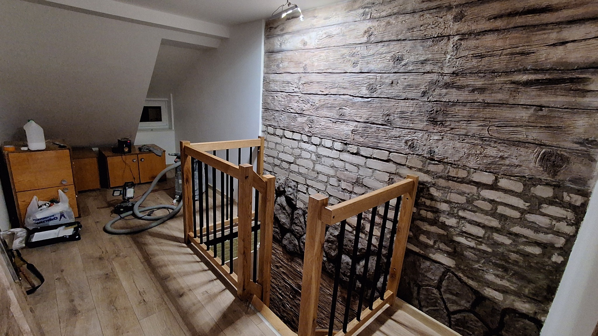 Schody z drewnianą balustradą i ozdobną fototapetą imitującą starą cegłę i drewno, widok z góry, na podłodze panele, w tle szafki i sprzęt.