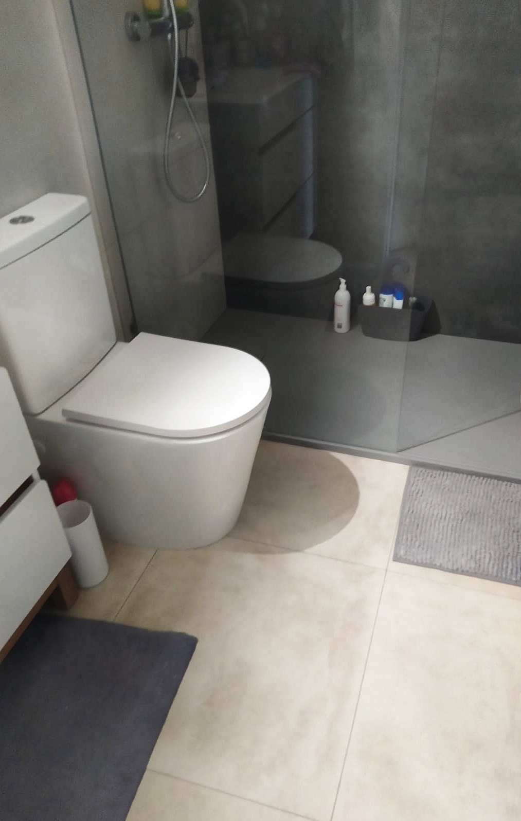Nowoczesna toaleta z bezkołnierzową miską WC i kabiną prysznicową typu walk-in z szarymi płytkami w tle, uzupełniona o beżowe płytki podłogowe i szarą wycieraczkę.