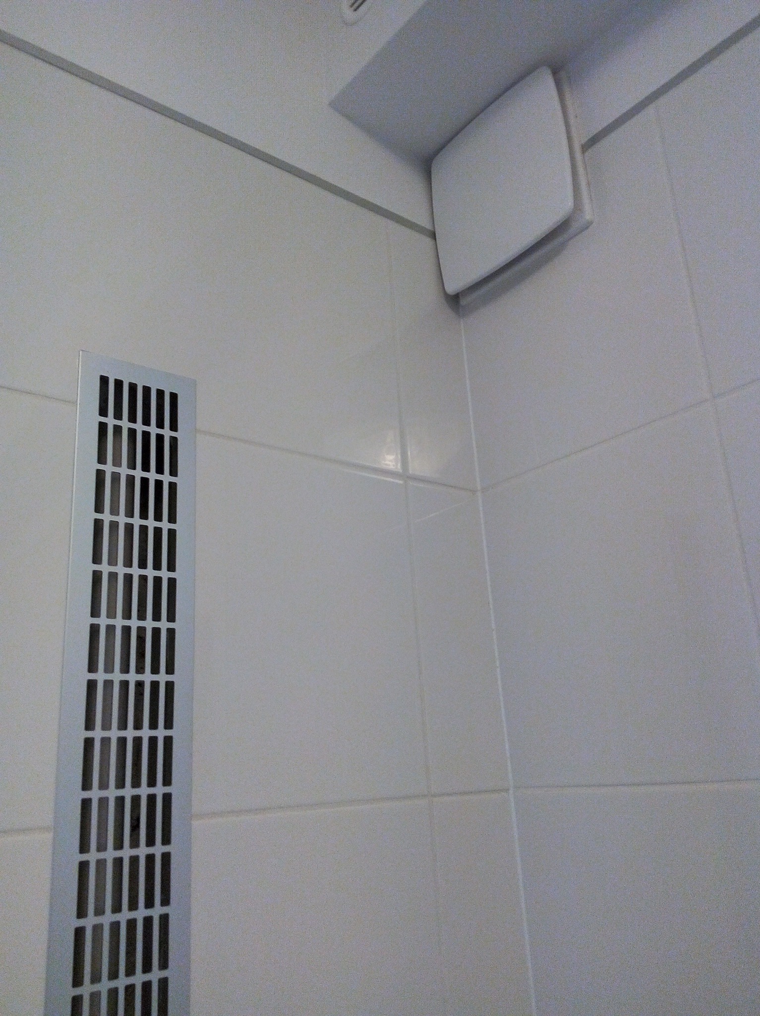 Szczegół ściany wyłożonej białymi płytkami z widoczną kratką wentylacyjną i wentylatorem sufitowym, ujęcie z dołu.