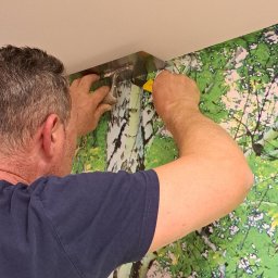 Budomex Sp. z o.o. - Pracownik dociska tapetę z motywem drzew do ściany za pomocą metalowej szpachelki i nożyka przy suficie.