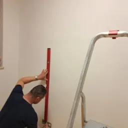 Elektryk z zegarkiem na ręce, przy użyciu poziomicy i ołówka, zaznacza punkty na ścianie nad gniazdkiem elektrycznym; drabina aluminiowa obok.