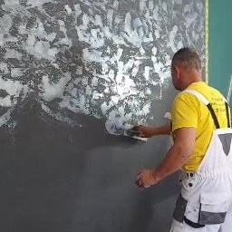 Budomex Sp. z o.o. - Nakładanie dekoracyjnej, srebrzystej farby na ciemną ścianę przez malarza w białych spodniach roboczych i żółtej koszulce. Efektowna faktura na ścianie.