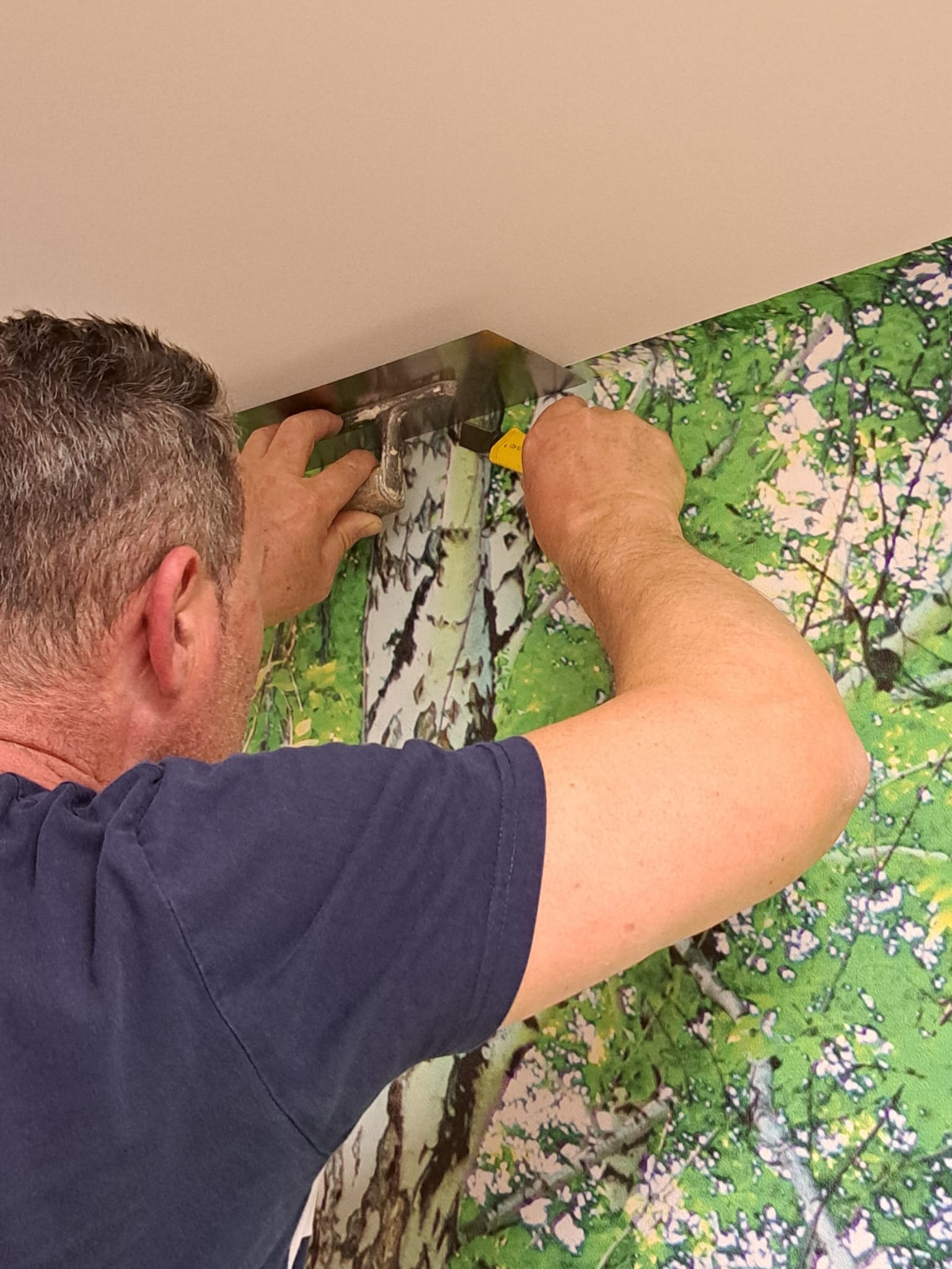 Pracownik dociska tapetę z motywem drzew do ściany za pomocą metalowej szpachelki i nożyka przy suficie.