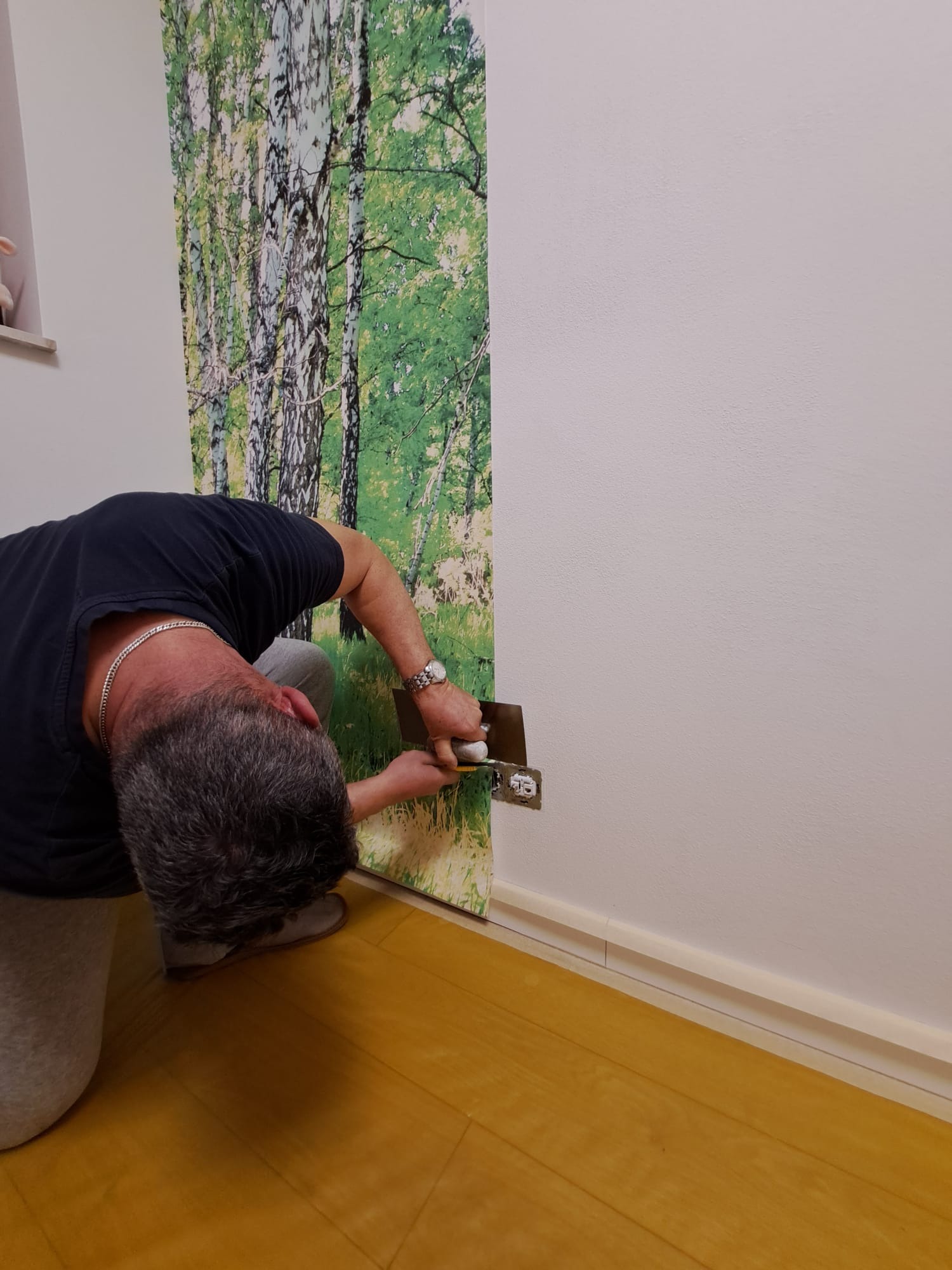 Mężczyzna w trakcie tapetowania ściany z fototapetą imitującą las brzozowy, docinanie tapety wokół gniazdka elektrycznego, widoczne narzędzia i fragment listwy przypodłogowej.
