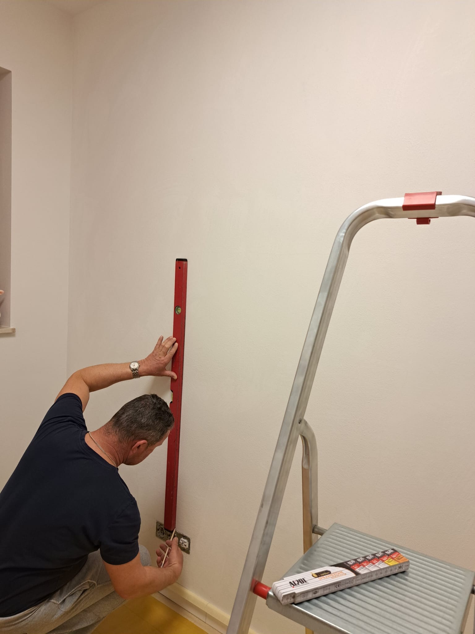 Elektryk z zegarkiem na ręce, przy użyciu poziomicy i ołówka, zaznacza punkty na ścianie nad gniazdkiem elektrycznym; drabina aluminiowa obok.