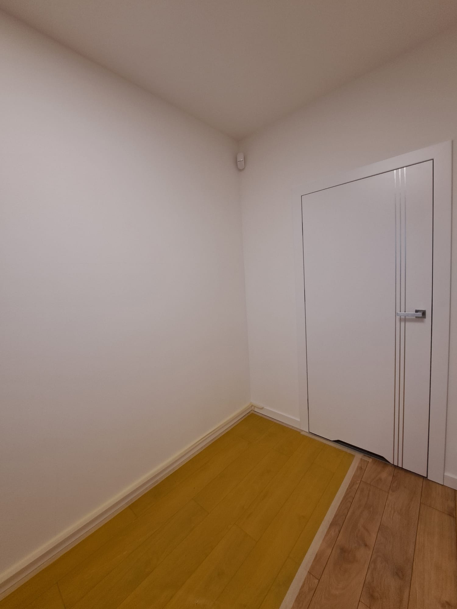 Wnętrze z białą ścianą, drzwiami z trzema pionowymi paskami i podłogą z dwoma rodzajami paneli: żółtymi i drewnopodobnymi.