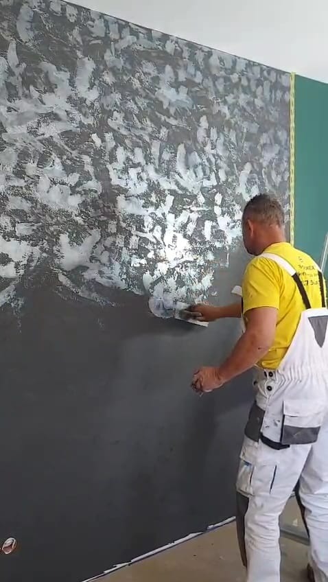 Nakładanie dekoracyjnej, srebrzystej farby na ciemną ścianę przez malarza w białych spodniach roboczych i żółtej koszulce. Efektowna faktura na ścianie.