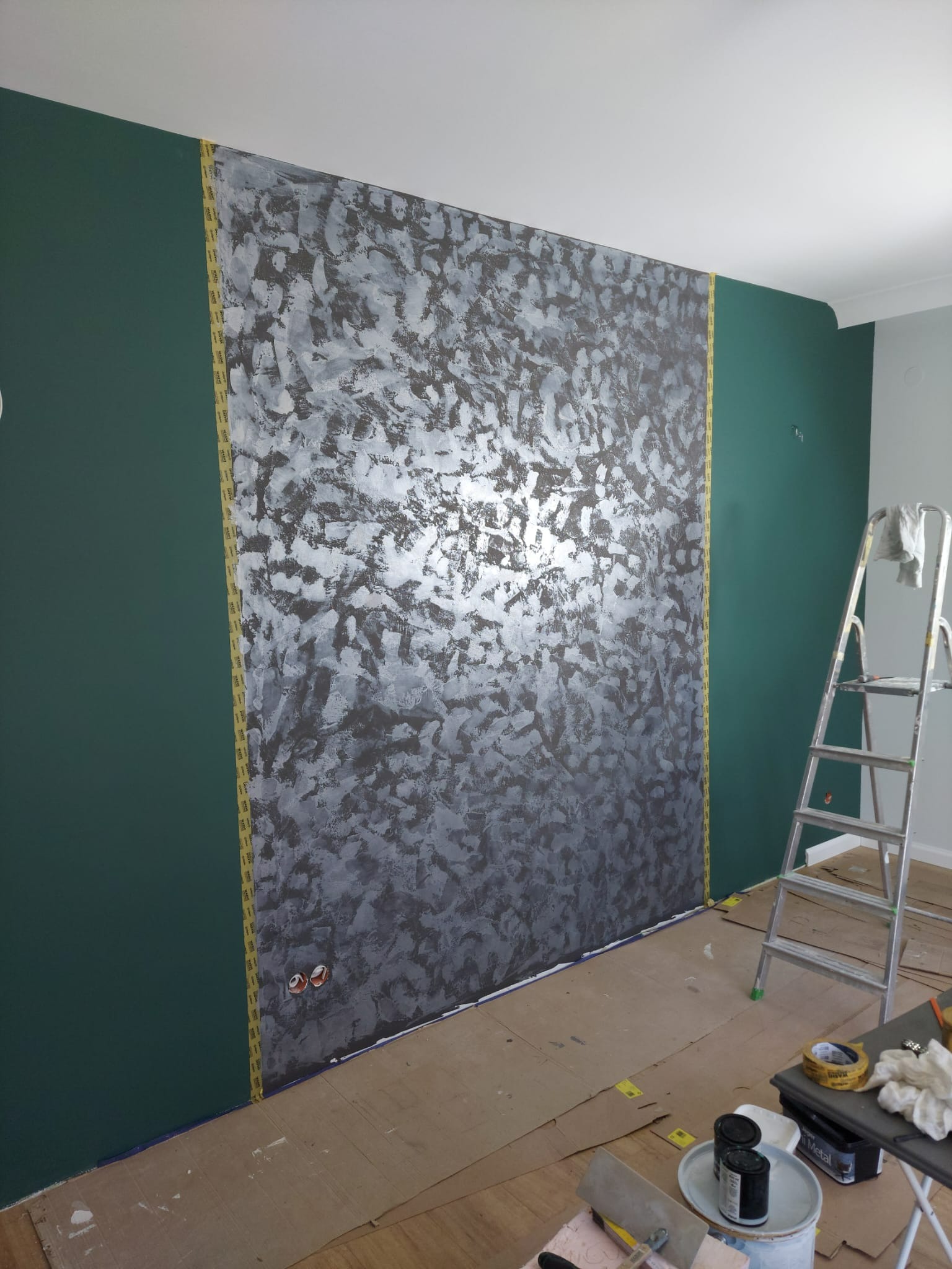 Ściana z dekoracyjną strukturą w odcieniach szarości, oddzielona taśmą malarską od zielonej ściany. Widoczne narzędzia malarskie i drabina w pomieszczeniu.